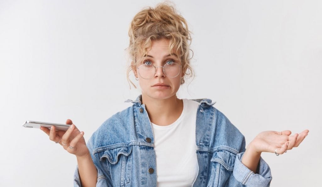 confused woman with glasses on, a jean shirt and hair in a bun holding a smartphone in her right hand