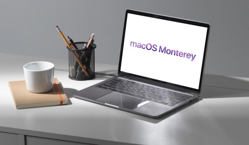 MacBook com MacOS Monterey numa secretária com suporte de caneta, um caderno e uma caneca