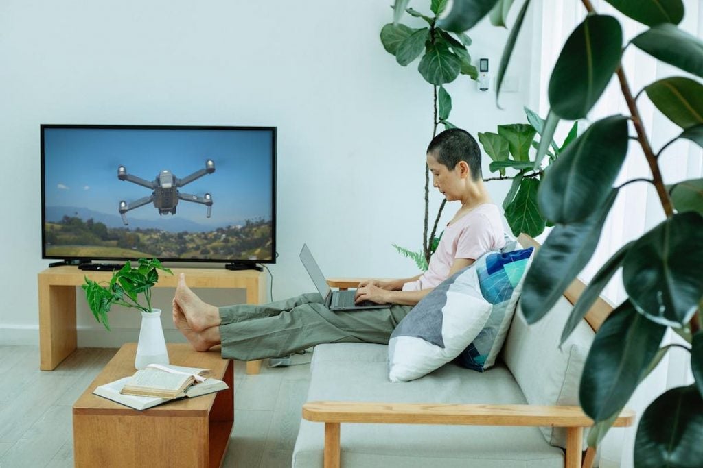 a man sitting on a couch with his feet on the table. There is a laptop on his lap. There is a Smart Tv hanging on a wall opposite of the man. the Smart TV displays an image of a drone