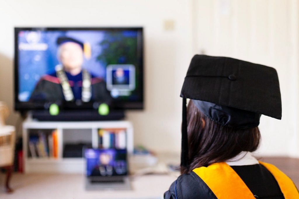 une femme en tenue de diplômée et coiffée d'une casquette voit l'image d'un homme en tenue d'avocat se refléter d'un ordinateur portable sur une télévision.