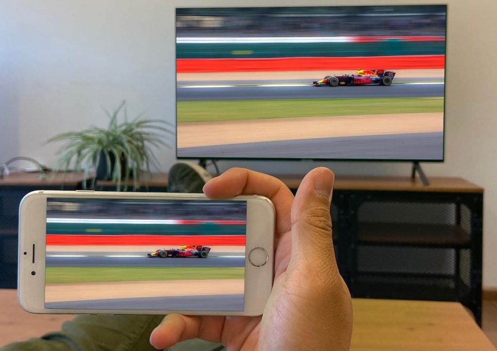 a person's hand holding an iPhone. the iPhone is mirroring Formula 1 to a TV standing on a short shelving system, with a plant to the left. The person's legs are crossed on the table.
