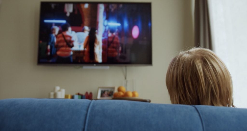 um topo da cabeça de uma pessoa é visível por detrás de um sofá. A pessoa está a olhar para uma televisão pendurada na parede