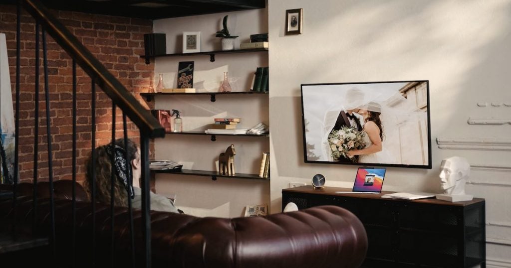 woman sitting on a black couch. She has a black bandana in her hair. She watches TV that displays an image of a kissing wedding couple mirrored from a laptop