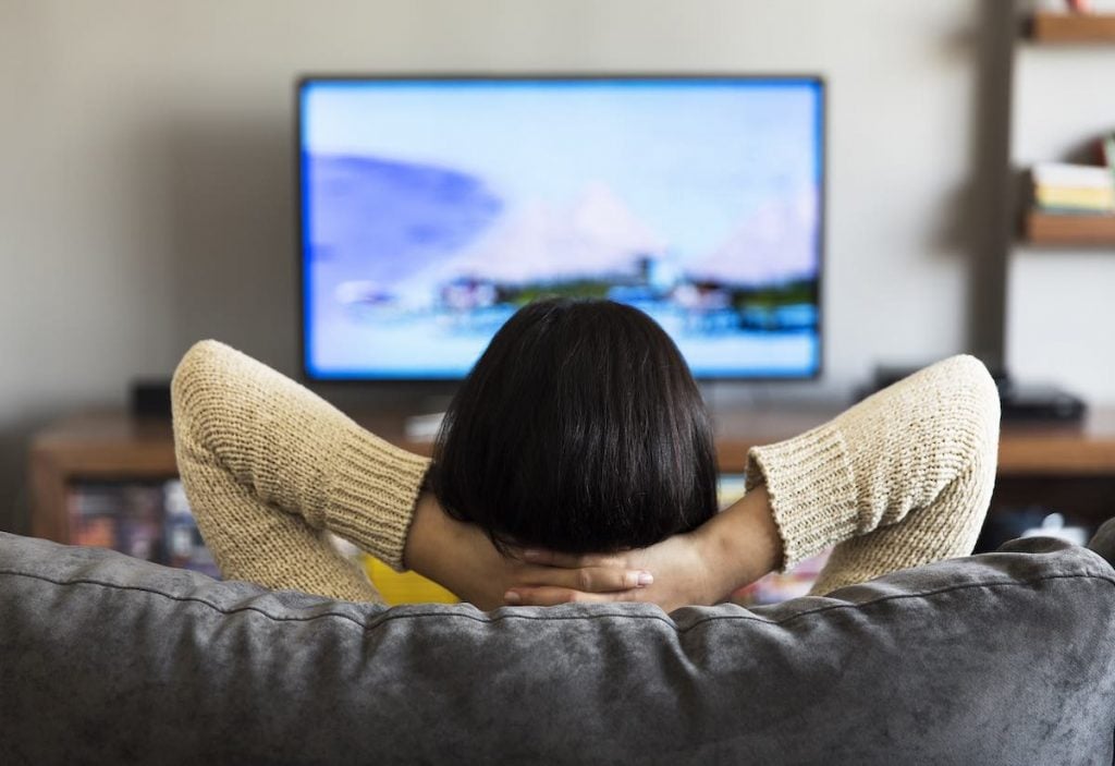 a woman sitting on a couch with her hands resting at the back of her neck. She is watching TV