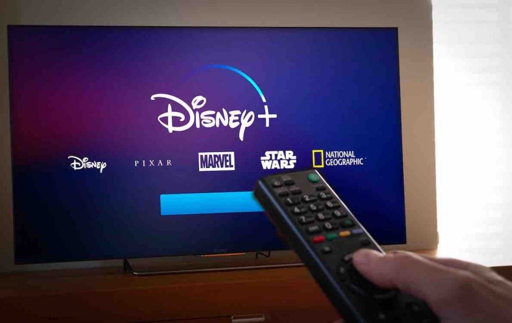 a hand holding a remote that's pointed at a Smart Tv. The Smart TV displays a disney plus logo