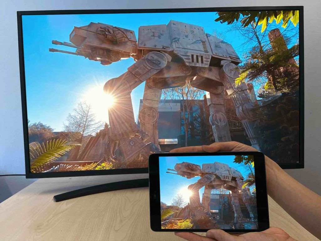 a hand holding an iPad. the iPad is casting an image of ATAT from Star Wars to a Smart TV. The Smart TV is resting on a table