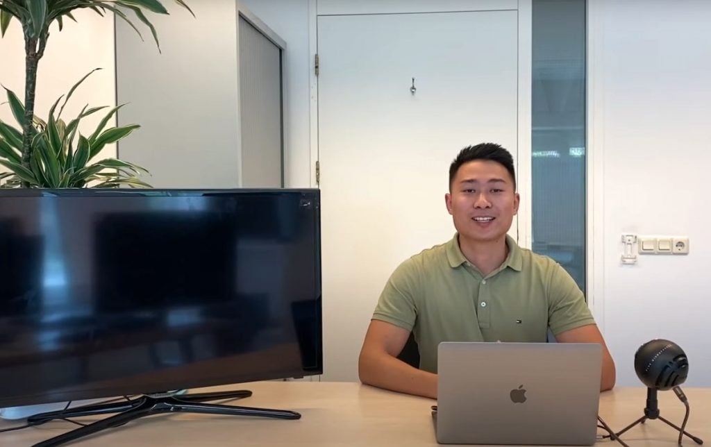 An Asian man is sitting at a table with a MacBook in front of him. There is a microphone and a Smart TV to his sides. There is a large houseplant in the room he's in