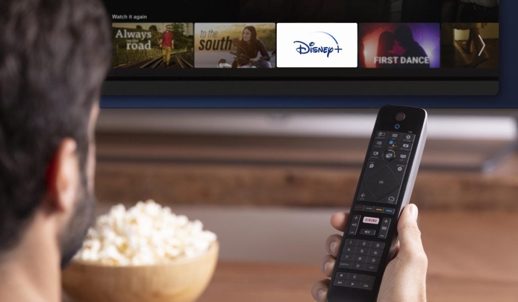 Een man houdt een afstandsbediening vast. Voor hem staat een schaal popcorn. De man zit voor een TV scherm met het Disney Plus logo erop.
