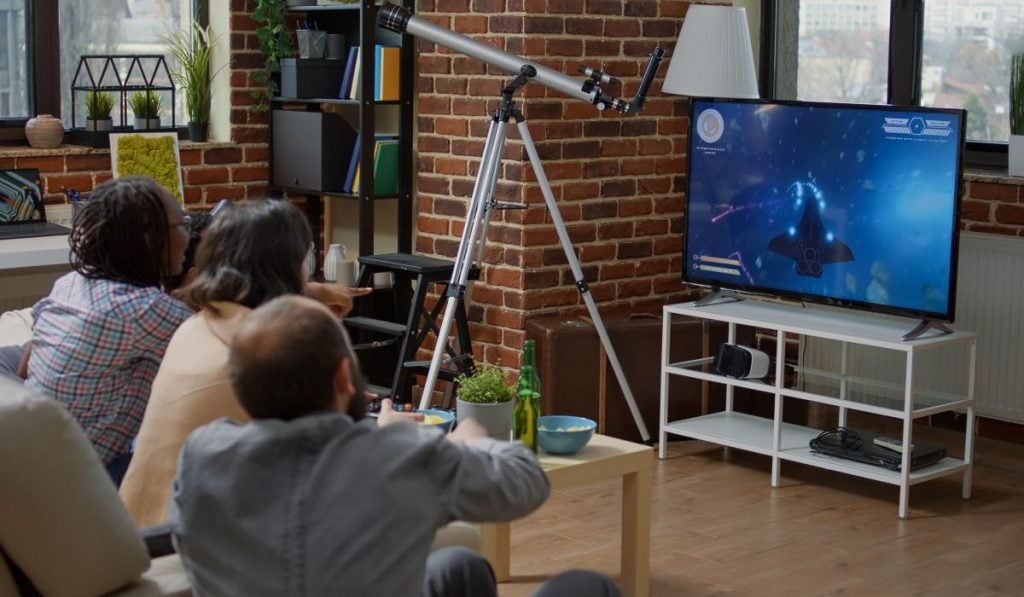 three people sitting on a couch watching TV. There's a telescope next to the TV