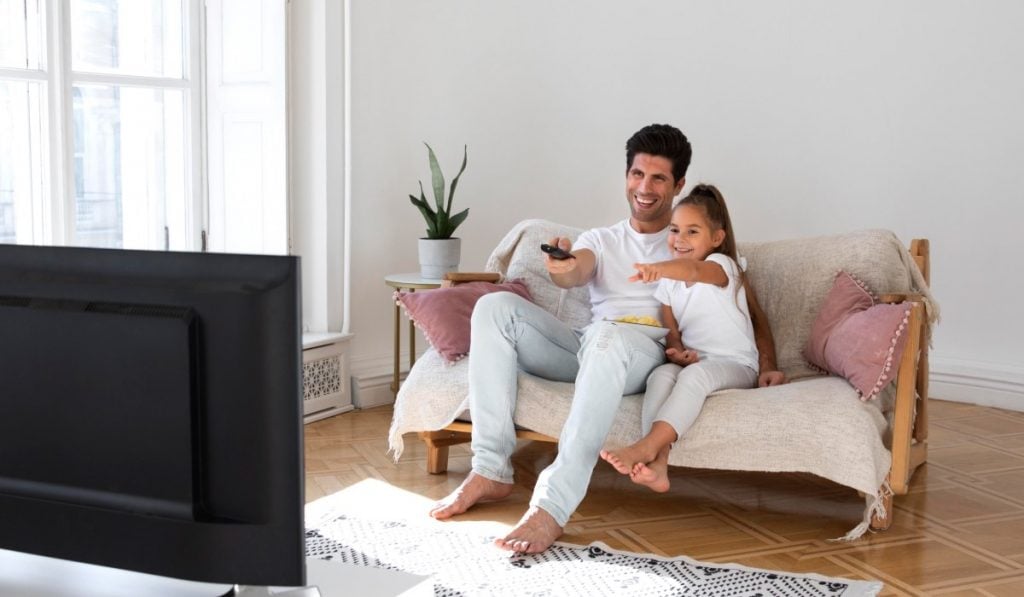 Een volwassen man en een kind zitten op een bank met een schaal snacks. De man houdt een afstandsbediening vast, terwijl het kind naar het tv-scherm wijst. Voor de man en het kind staat een televisie.