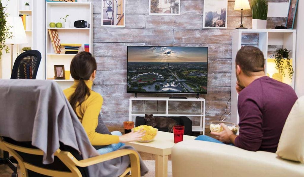 A man and a woman sit on comfortable chairs. They are watching a Smart TV. The Smart Tv stands on a flimsy TV stand and is set against a grey brick wall