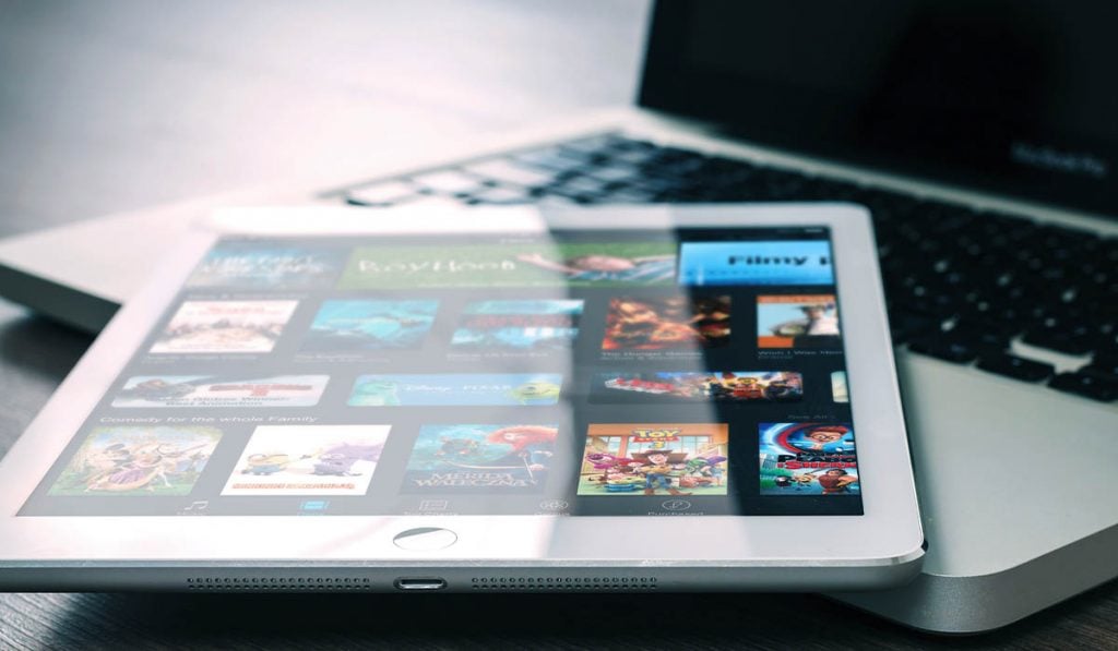 a tablet with a streaming service interface on the screen is laying on top of a laptop keyboard