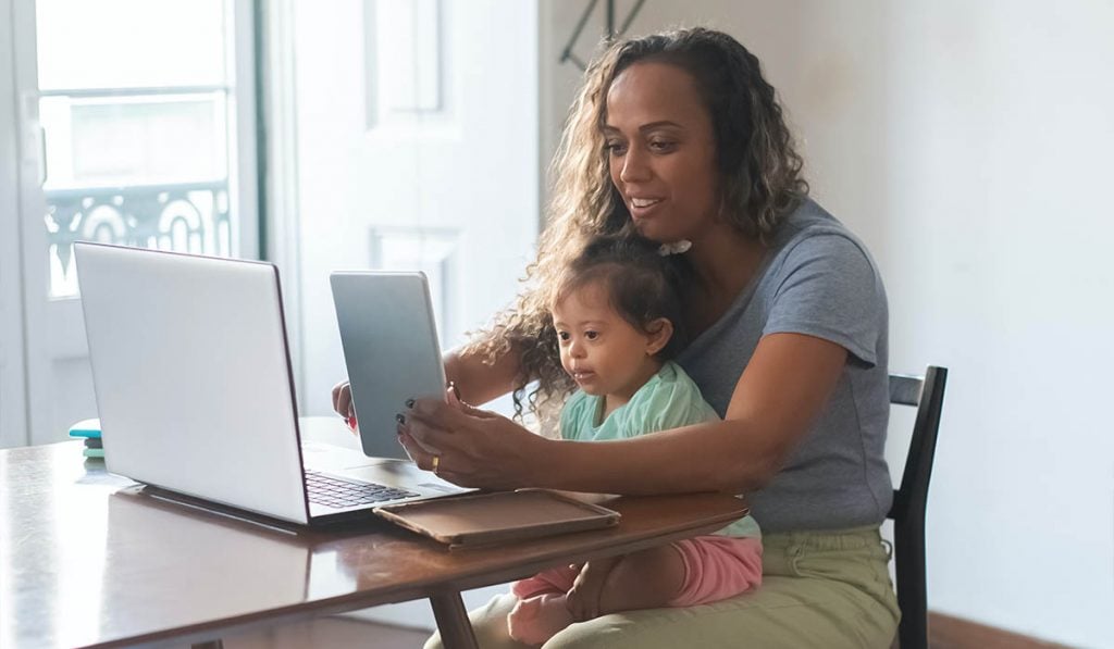 A woman with a baby on her lap is sitting by a table. There is a tablet in her hands and a laptop on the table.