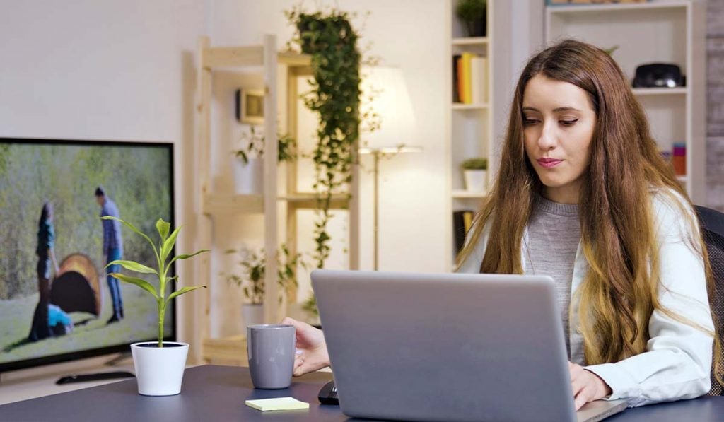 Woman sits at a table. She is holding a mug in one hand. The woman is using a laptop computer. There are shelves and a glassed cupboard behind her
