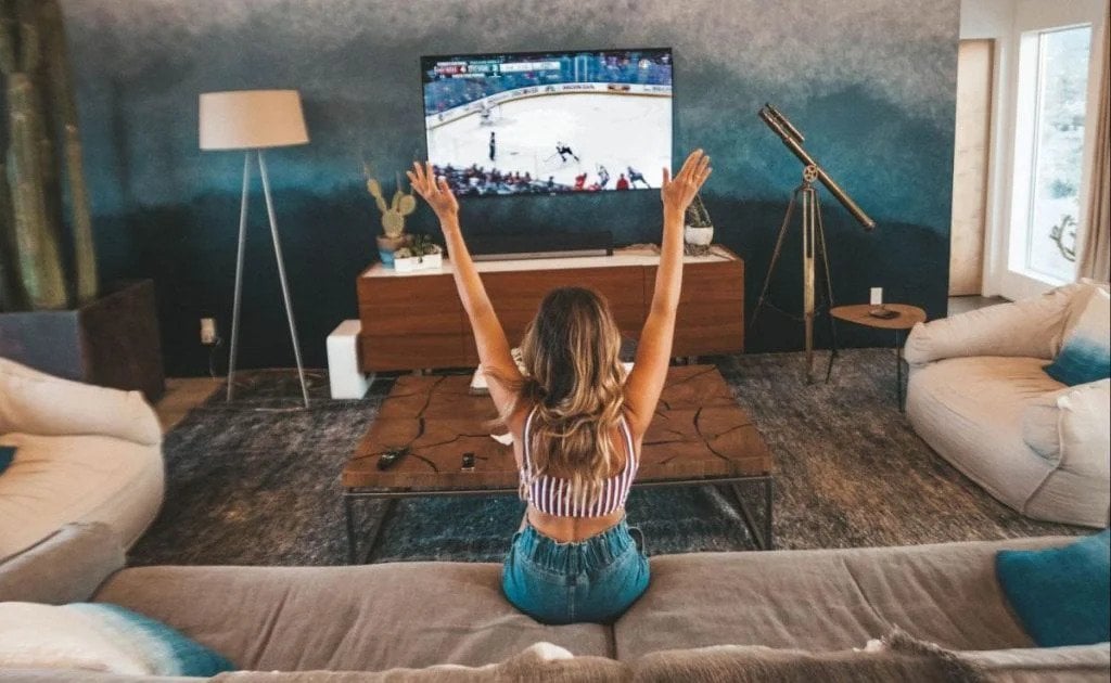 Woman sitting on a couch with her hair let down. She's watching sports on TV and cheering with her arms pointed upwards. The TV stands on a TV stand and there's a dark wall behind it. There's a lamp next to the TV
