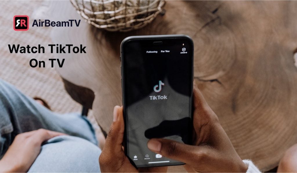 a person watching tik tok on a smartphone sitting next to a table made out of a slab of wood. The person is wearing blue jeans and there's a decorative basket on the table. In the top-right corner there's a header: 'Watch TikTok on TV' and the AirBeamTV logo