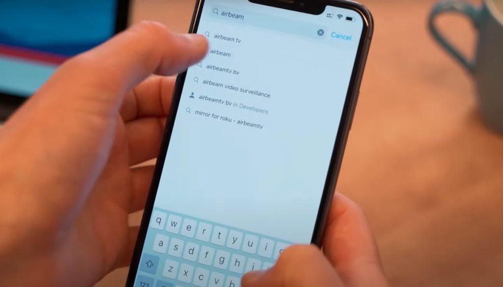 Two hands holding an iPhone. The iPhone has a search tab open