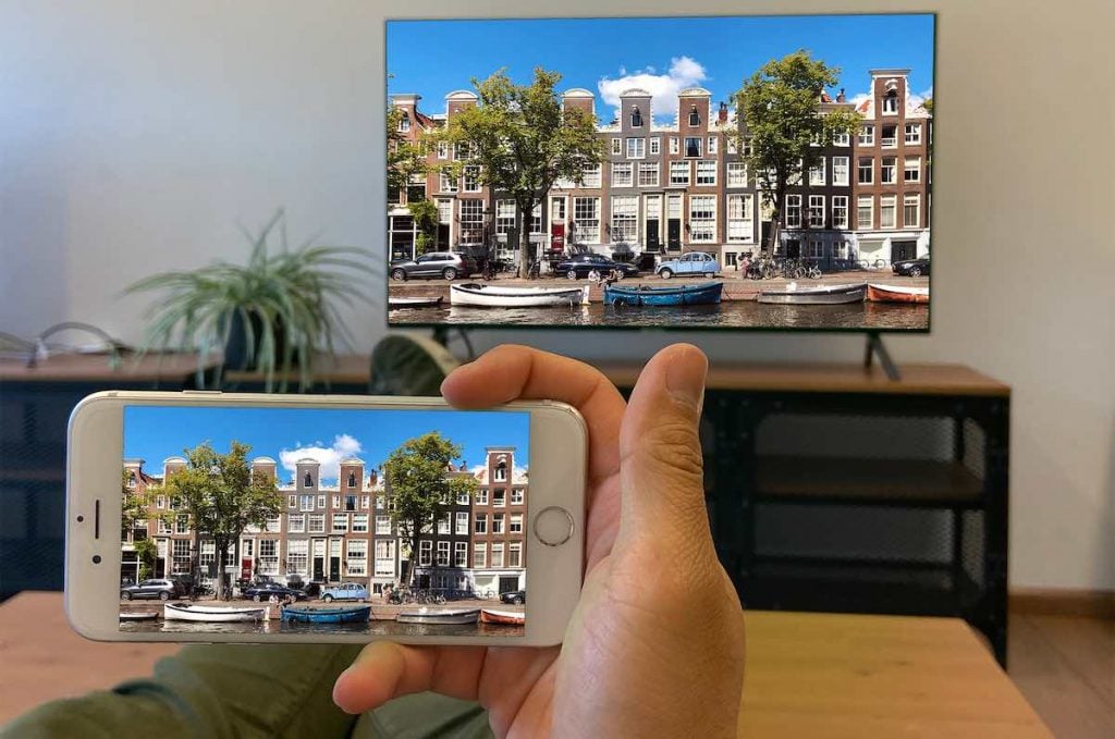 a person mirroring an image of Amsterdam from an iPhone to a Smart TV. The person has their legs on a table. The Smart TV stands on a shelving system with a plant to the right.