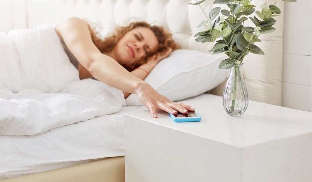 Woman laying in bed with an unhappy face. She's grabbing an iPhone that's resting on the night stand, next to a vase of roses