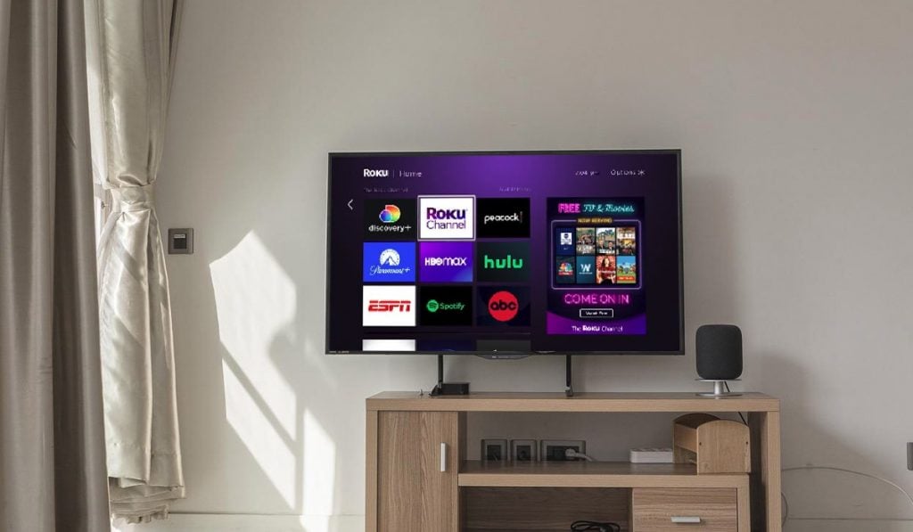 Roku interface on a Smart TV. There are multiple app icons on the TV screen including  Hulu, HBO Max, Spotify and Peacock. the Smart TV is located in a TV stand next to a window with curtains