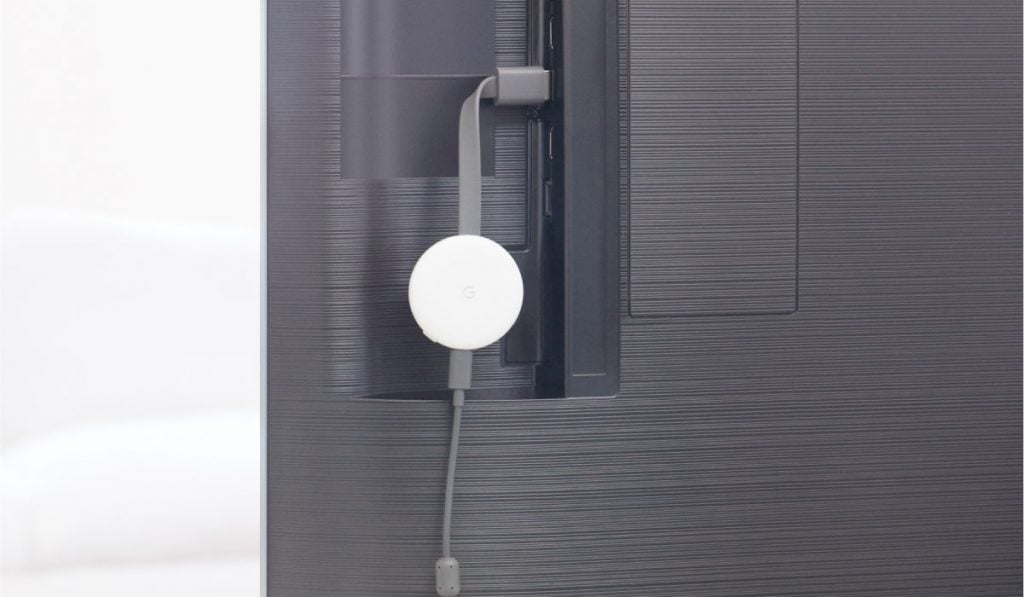A chromecast device plugged into the back of a Smart TV through an HDMI plug. The Chromecast is plugged to a power cable