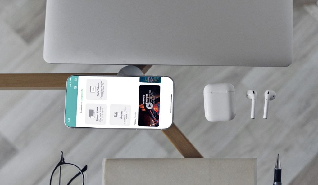 an iPhone with the Mirorr for LG TV app open laying on a glass table. The iPhone is surrounded by a pair of AirPods, an AirPod case, a MacBook, a pair of glasses, a pen and a sachet. White wooden floor is visible from under the table