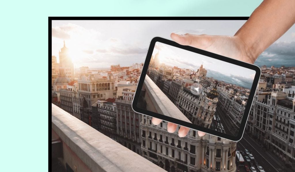 a person holding an iPad. The iPad mirrors an image of an old city during the sunset to a screen
