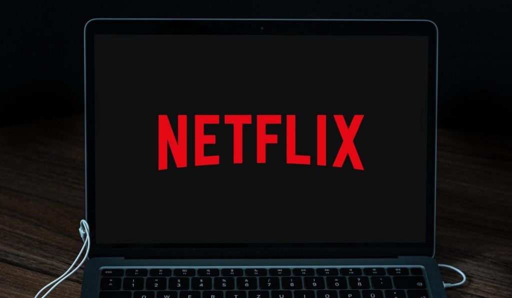 a laptop in a darkened room with a pair of earphones and a charging wire plugged in. The laptop screen displays Netflix logo on black background. The laptop rests on wooden surface