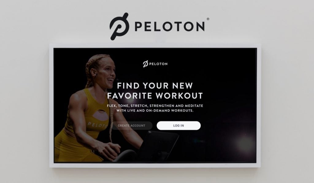 A Peloton landing page with a slogan and two action buttons overlaying an image of a smiling woman using a training bike