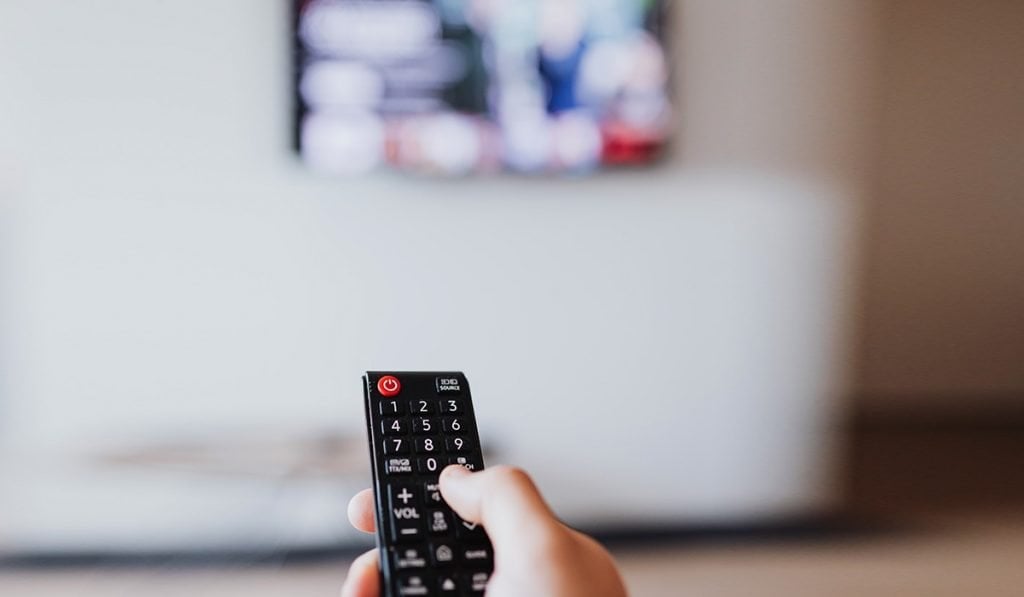 Una mano che tiene un telecomando e lo punta verso un televisore. La TV è sfocata e il telecomando è a fuoco.