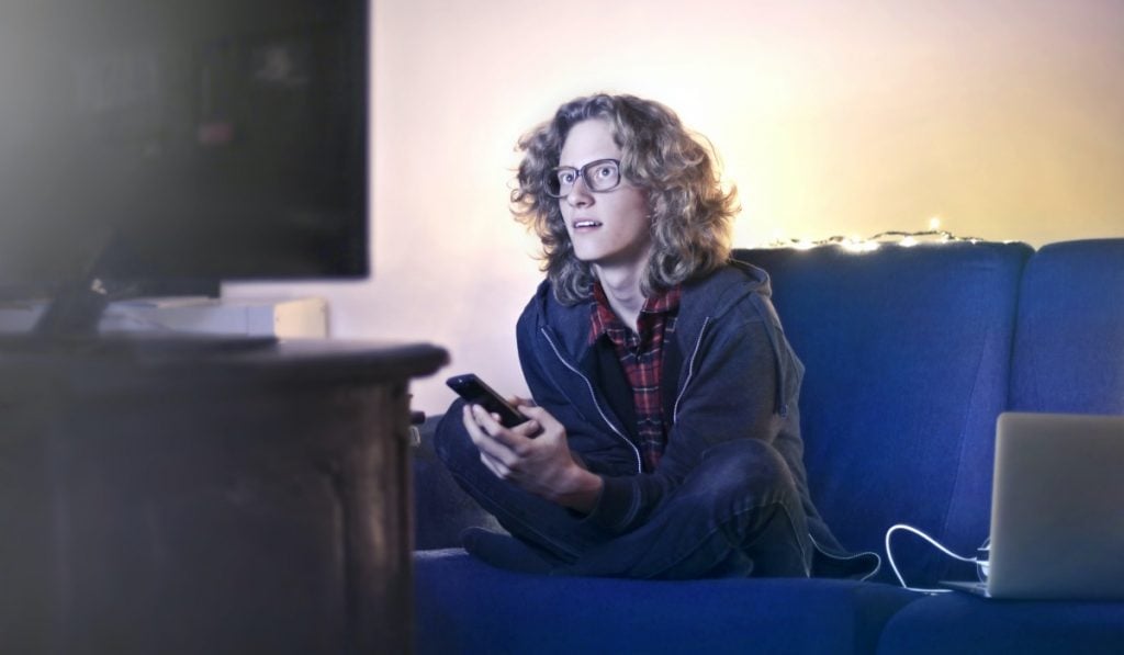 Persona con gli occhiali seduta a gambe incrociate su un divano. la persona tiene un telefono in mano. c'è una TV di fronte alla persona e la persona sta guardando la TV con il viso concentrato.