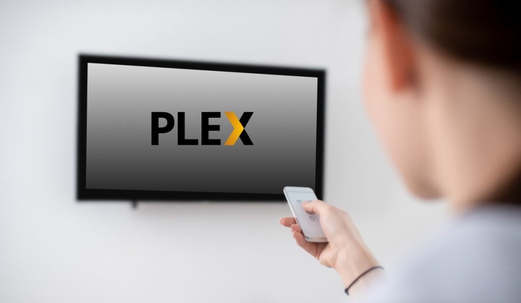 a person using a TV remote to use a Smart TV that's hanging on a wall. The Smart TV has the Plex logo on the screen