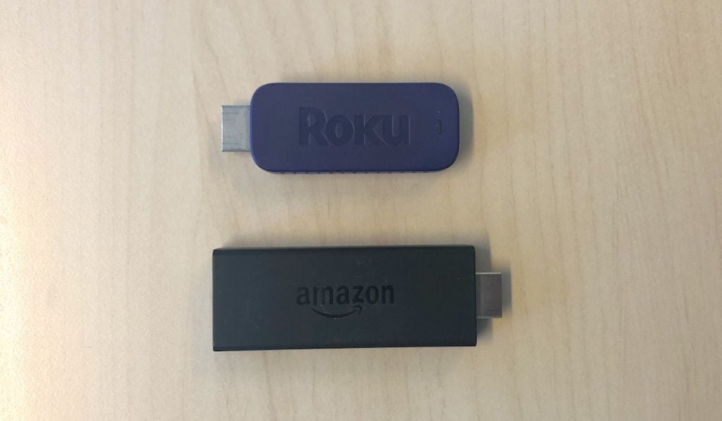 Roku stick and Fire TV Stick laying on a wooden surface in paralell