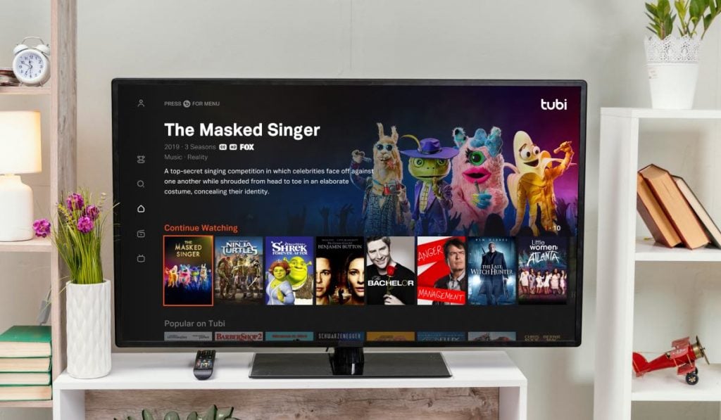 a Smart TV standing on a TV stand surorunded by shelves with lamps, plants, trinkets and books. The TV screen displays the Tubi TV interface with The Masked Singer film page on