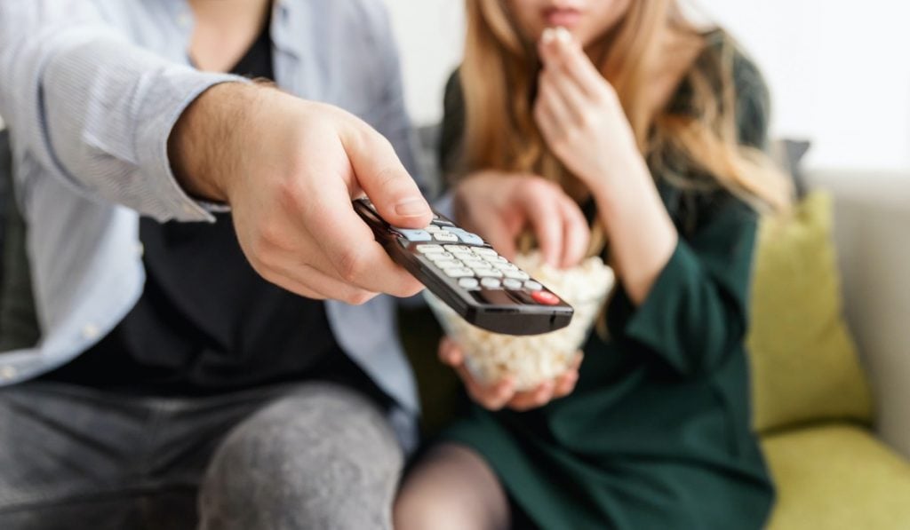 two people sit on a couch. One of them is holding a remote and pointing it forwards while pressing a button. The other sits back and eats popcorn out of a glass bowl