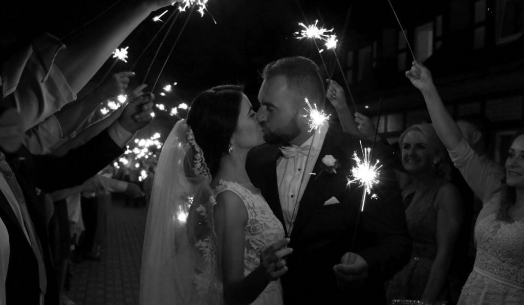 zwart-wit foto van een pasgetrouwd stel dat zoent en omringd wordt door gasten die vuurwerk vasthouden