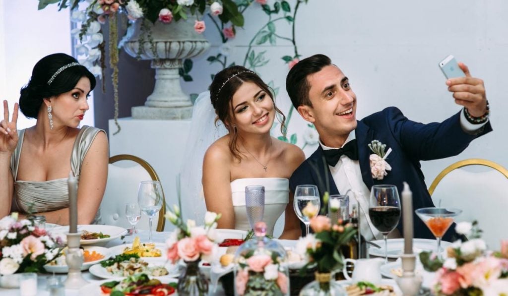 twee vrouwen en een man zitten aan een bruiloftsfeesttafel en nemen een selfie