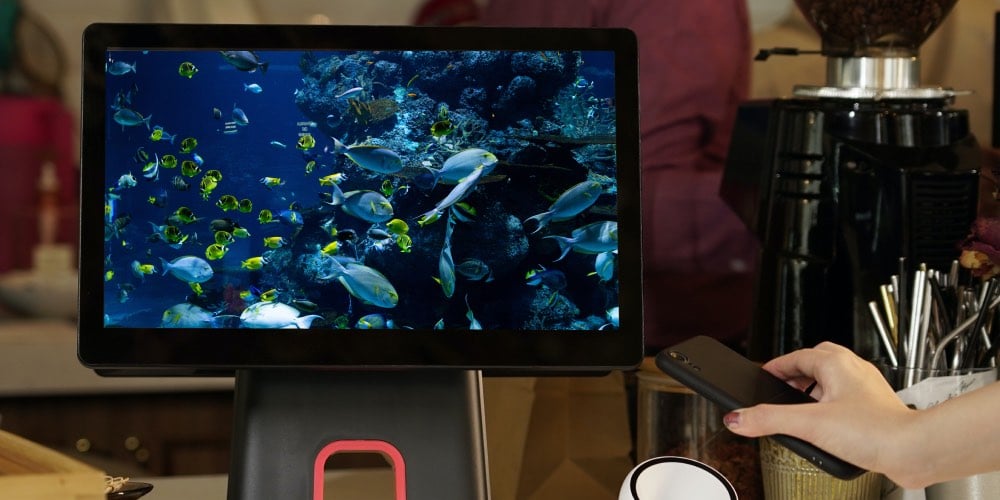 een hand met gelakte nagels die een smartphone vasthoudt, een schermpaneel met vissen rond een stuk koraalrif en een koffiezetapparaat op de achtergrond