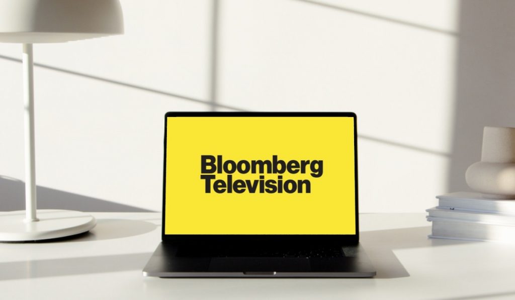 bloomberg TV logo ơn a laptop standing on a desk with a desk lamp and some books