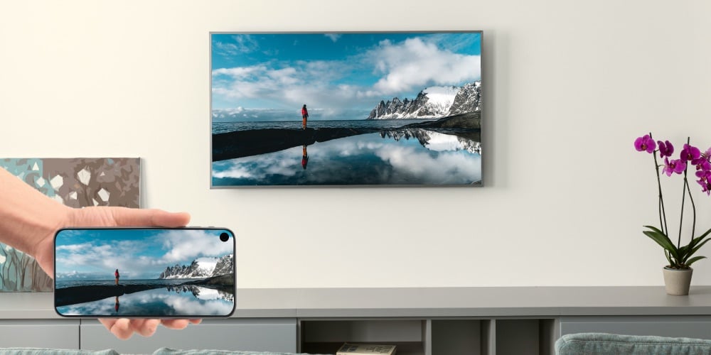 person holding a smartphone casting an image of a person standing at the shore of a lake with snowy mountains and clouds in the background. The image is being casted to a Samsung TV that hangs on a wall above a low cupboard with some flowers in a pot