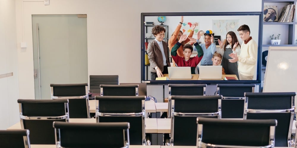 een klaslokaal vol zwarte stoelen. aan de muur hangt een Smart TV met een beeld van juichende kinderen naast hun leraar. De kinderen gebruiken Apple laptops