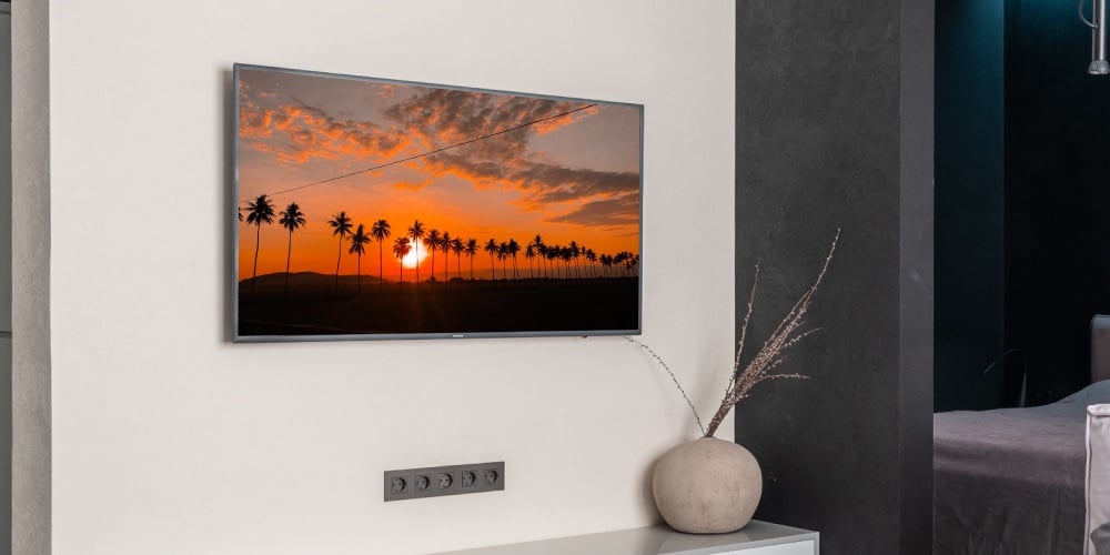 an image of palms against a sunset displayed on TV hanging on a wall above a wall-mounted power socket. there's a decorative element in the bottom-middle of the image