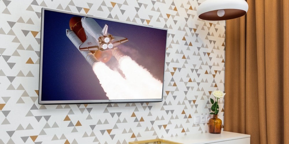 een beeld van een ruimte-shuttle dat de ruimte in vliegt wordt getoond op een smart tv. de smart tv hangt aan een muur met een behang met kleurrijke driehoeken. aan de rechterkant van het beeld is een gordijn getekend.