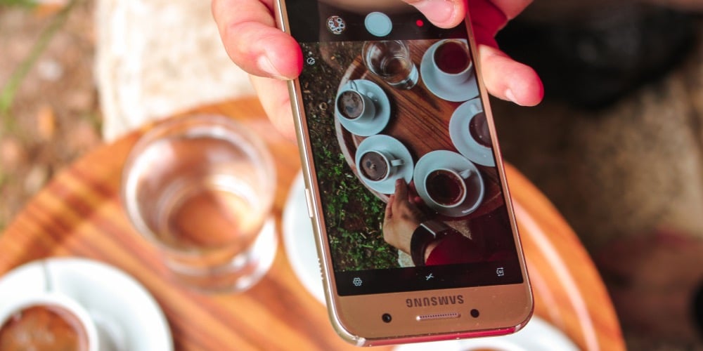 a person holding a samsung smartphone taking a photo of a round wooden table with several cups of coffee