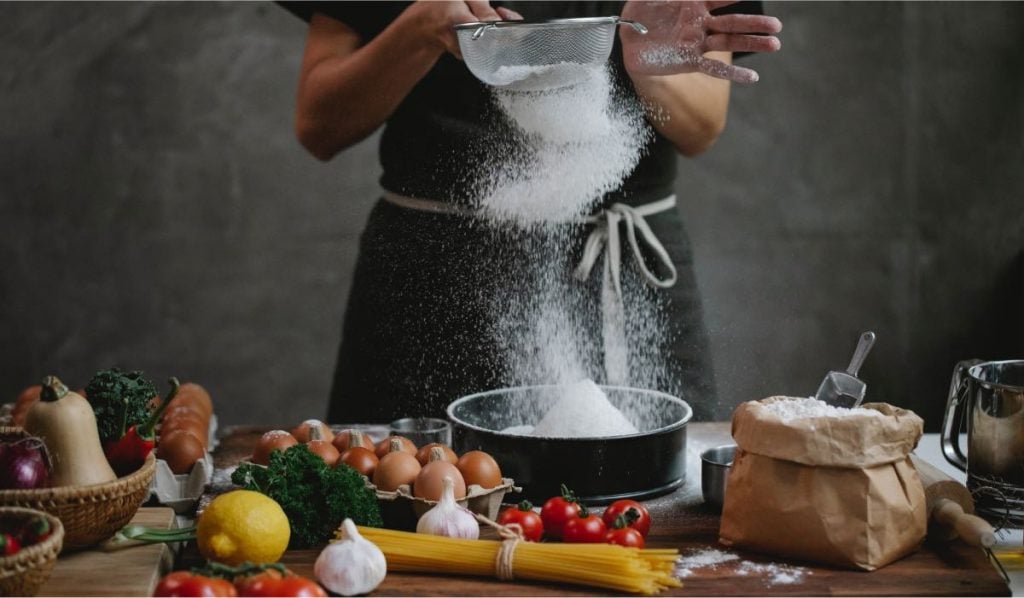woman sifting flour in a kitchen on a table with various vegetables