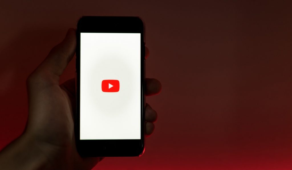 una mano che tiene uno smartphone con il logo di YouTube su sfondo bianco