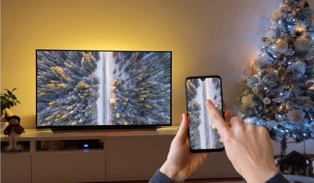 a hand holding a smartphone and the other hand pointing at a screen. The smartphone casts an image of a birds-eye view of a forest and a road cutting through it. There is a Christmas tree on the right side of the image