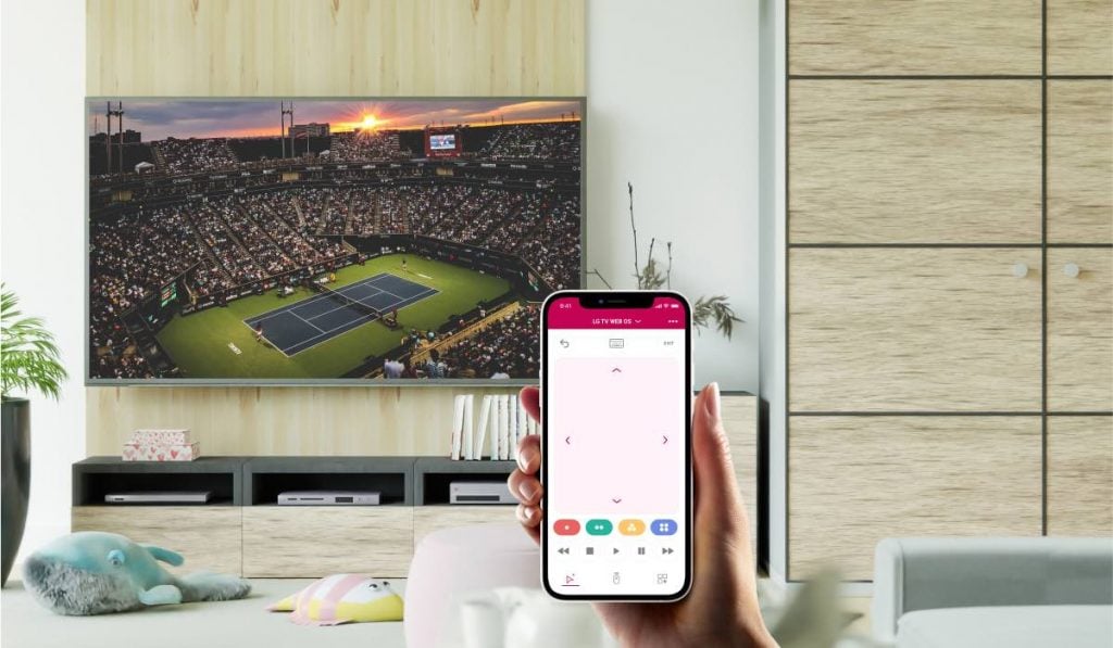 A hand holding an iPhone with the LG Tv remote app by MeisterApps on the screen. In the living room, there is a large wardrobe, and a wooden wall, upon which an LG TV is hanging. There are two stuffed toys on the flooe