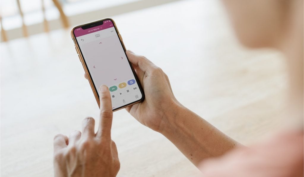 a shoulder view of a person holding an iPhone with one hand and operating it with the pointing finger of the other hand. The iPhone screen has the LG TV remote app interface on the screen.