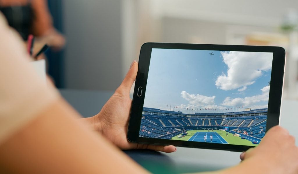 A person using a tablet, propping it up with their arms. The tablet displays an image of an empty stadium
